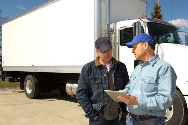 A fleet owner stands next to one of his employees and a truck, showing real-time fleet card analytics on his tablet.